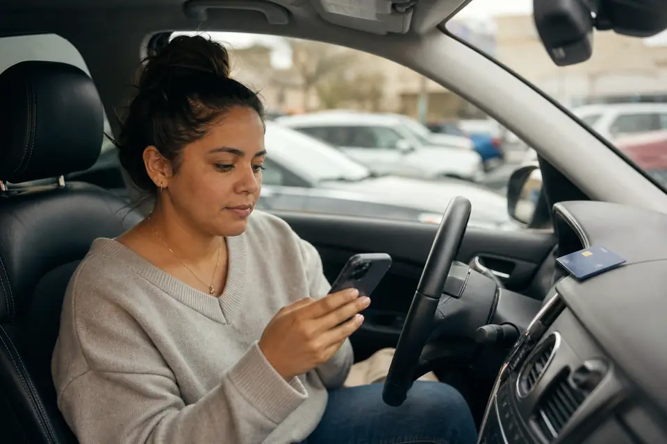 Mujer revisa su teléfono en el auto, tarjeta sobre el tablero.